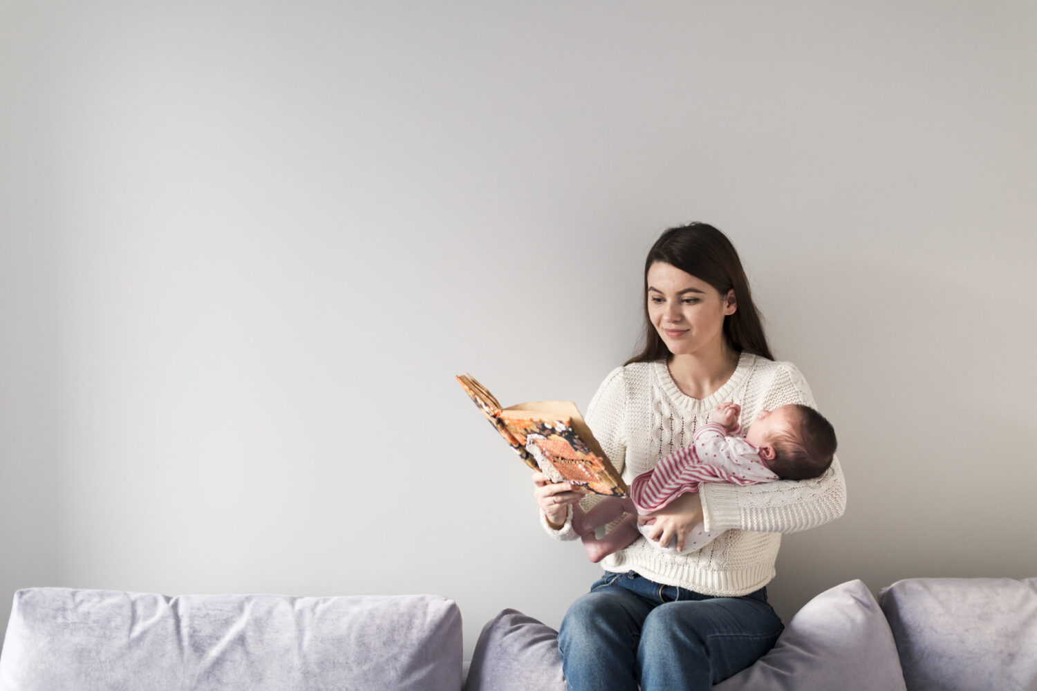 Mother singing a lullaby from book to her baby on her lap in the bedroom