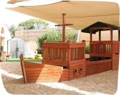 Wooden pirate-themed playground at Step by Step Nursery in Dubailand, featuring a ship-like structure with a mast and shaded sandy play area.